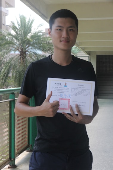 A person is standing in a corridor holding a diploma or certificate. They are wearing a black shirt and are giving a thumbs-up gesture. The background includes a palm tree and part of the building structure.