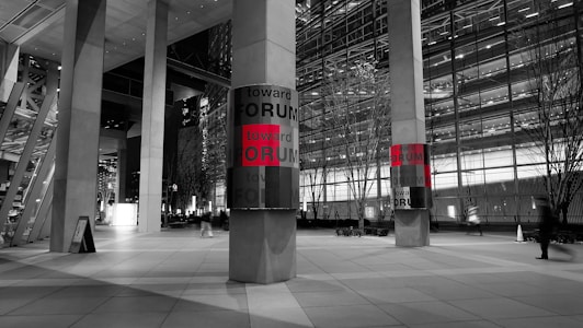 A modern urban setting displays two large vertical columns with panels that read 'toward FORUM' in bold letters, with the word 'FORUM' highlighted in red. The scene is predominantly monochrome, with the red text offering a striking contrast. Tall buildings with glass facades and sparse trees line the background, while a few blurred figures suggest a bustling yet serene cityscape at night.