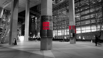 A modern urban setting displays two large vertical columns with panels that read 'toward FORUM' in bold letters, with the word 'FORUM' highlighted in red. The scene is predominantly monochrome, with the red text offering a striking contrast. Tall buildings with glass facades and sparse trees line the background, while a few blurred figures suggest a bustling yet serene cityscape at night.