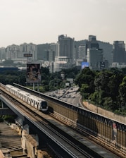 A sleek train travels on an elevated track through a cityscape, surrounded by lush greenery. Tall skyscrapers and buildings are visible in the background, partially shrouded by a hazy sky. A highway with moving cars runs parallel to the train track, adding to the urban setting.