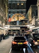 A bustling evening street scene in front of a luxurious hotel entrance adorned with ornate decorations. Cars, including a black BMW, are parked along a paved driveway lined with people walking or standing. The hotel features an elegant facade with illuminated signage and festive decorations, including Christmas trees and lights.