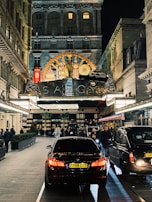A bustling evening street scene in front of a luxurious hotel entrance adorned with ornate decorations. Cars, including a black BMW, are parked along a paved driveway lined with people walking or standing. The hotel features an elegant facade with illuminated signage and festive decorations, including Christmas trees and lights.
