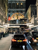 A bustling evening street scene in front of a luxurious hotel entrance adorned with ornate decorations. Cars, including a black BMW, are parked along a paved driveway lined with people walking or standing. The hotel features an elegant facade with illuminated signage and festive decorations, including Christmas trees and lights.