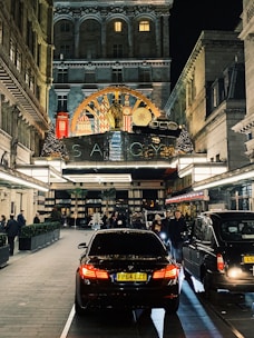 A bustling evening street scene in front of a luxurious hotel entrance adorned with ornate decorations. Cars, including a black BMW, are parked along a paved driveway lined with people walking or standing. The hotel features an elegant facade with illuminated signage and festive decorations, including Christmas trees and lights.