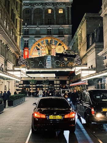 A bustling evening street scene in front of a luxurious hotel entrance adorned with ornate decorations. Cars, including a black BMW, are parked along a paved driveway lined with people walking or standing. The hotel features an elegant facade with illuminated signage and festive decorations, including Christmas trees and lights.