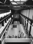 An expansive indoor space with a high ceiling, featuring industrial-style architecture. People are scattered across the open floor, some standing in groups and others moving throughout the area. The space has metal beams and railing, lending a modern and minimalistic feel.
