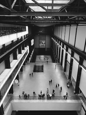 An expansive indoor space with a high ceiling, featuring industrial-style architecture. People are scattered across the open floor, some standing in groups and others moving throughout the area. The space has metal beams and railing, lending a modern and minimalistic feel.
