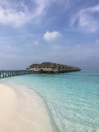 A row of overwater bungalows extends into a calm turquoise sea with a wooden boardwalk connecting them to the white sandy beach. The sky is clear with a few scattered clouds, contributing to the serene and peaceful atmosphere.