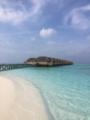 A row of overwater bungalows extends into a calm turquoise sea with a wooden boardwalk connecting them to the white sandy beach. The sky is clear with a few scattered clouds, contributing to the serene and peaceful atmosphere.