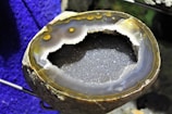 A geode with a rough outer surface and a sparkling, crystalline interior. The inner layers display various shades of gray and brown, with some areas showing a hint of yellow. The background is a vibrant blue, providing contrast to the earthy tones of the geode.