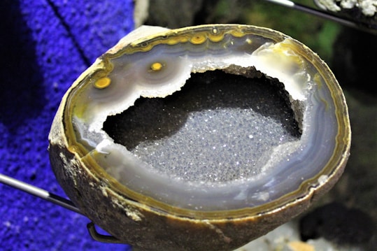 A geode with a rough outer surface and a sparkling, crystalline interior. The inner layers display various shades of gray and brown, with some areas showing a hint of yellow. The background is a vibrant blue, providing contrast to the earthy tones of the geode.