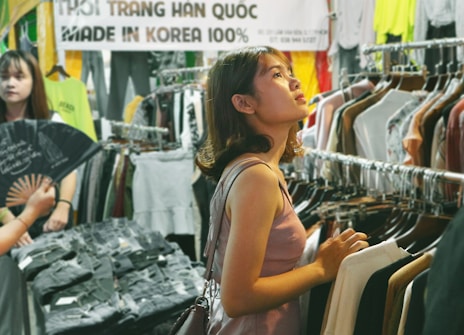 A young woman stands in a clothing store, surrounded by racks of various garments. She is gazing upwards, possibly considering her choices. Another person is nearby, partially visible and holding a fan. The store has a casual, vibrant atmosphere with a large sign in the background that reads 'Made in Korea 100%'.
