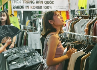 A young woman stands in a clothing store, surrounded by racks of various garments. She is gazing upwards, possibly considering her choices. Another person is nearby, partially visible and holding a fan. The store has a casual, vibrant atmosphere with a large sign in the background that reads 'Made in Korea 100%'.