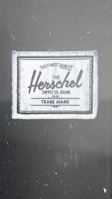A fabric label with the text 'The Finest Quality Herschel Supply Co. Brand Trade Mark'. The label has a vintage style with a rectangular shape, bordered by a dark line.