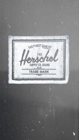 A fabric label with the text 'The Finest Quality Herschel Supply Co. Brand Trade Mark'. The label has a vintage style with a rectangular shape, bordered by a dark line.