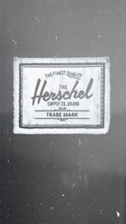 A fabric label with the text 'The Finest Quality Herschel Supply Co. Brand Trade Mark'. The label has a vintage style with a rectangular shape, bordered by a dark line.