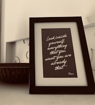 A framed quote is placed on a table. The quote reads, 'Look inside yourself; everything that you want, you are already that.' The frame has a sleek black border and sits next to a decorative bowl with a textured, dark surface. The background is neutral, contributing to a minimalist aesthetic.