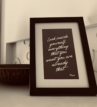 A framed quote is placed on a table. The quote reads, 'Look inside yourself; everything that you want, you are already that.' The frame has a sleek black border and sits next to a decorative bowl with a textured, dark surface. The background is neutral, contributing to a minimalist aesthetic.