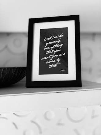 A black and white framed quote on a shelf, surrounded by a textured background. The quote reads, 'Look inside yourself; everything that you want, you are already that.' Attributed to Rumi, the text is written in a script font on a dark background.