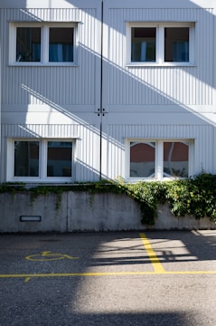 A two-story building with white panel siding is shown, featuring four windows with curtains. Green foliage grows over a concrete wall in front of the building. The ground is marked with a yellow handicap parking symbol and lines on the asphalt. Diagonal shadows from the building structure intersect the scene, creating a pattern on the surface.