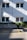 A two-story building with white panel siding is shown, featuring four windows with curtains. Green foliage grows over a concrete wall in front of the building. The ground is marked with a yellow handicap parking symbol and lines on the asphalt. Diagonal shadows from the building structure intersect the scene, creating a pattern on the surface.