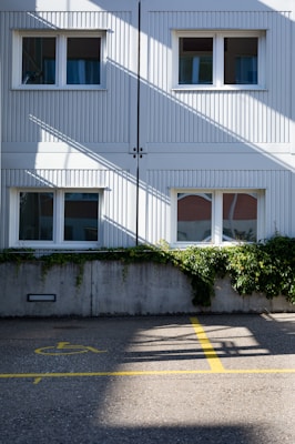 A two-story building with white panel siding is shown, featuring four windows with curtains. Green foliage grows over a concrete wall in front of the building. The ground is marked with a yellow handicap parking symbol and lines on the asphalt. Diagonal shadows from the building structure intersect the scene, creating a pattern on the surface.