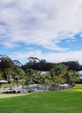 A lush, green lawn leads to a pool area bordered by palm trees. In the background, there are buildings with white roofs. The sky is expansive, filled with clouds and touches of blue.