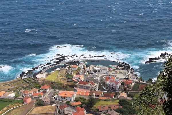 A coastal village with red-roofed buildings sits next to the ocean. The sea is vibrant blue with waves crashing against the shoreline, and there is a natural swimming pool formed by dark volcanic rocks. The area is lush with green vegetation surrounding the village.
