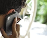 A person is holding a smartphone to their ear while wearing sunglasses. The background is blurred, showcasing outdoor greenery and a white metal structure.