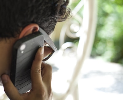A person is holding a smartphone to their ear while wearing sunglasses. The background is blurred, showcasing outdoor greenery and a white metal structure.