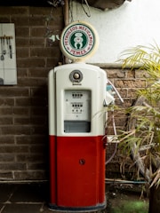 A vintage PEMEX gas pump is situated against a brick wall, partially covered by a palm plant. The pump is red and white, featuring the PEMEX logo on top. Beside it, a decorative panel displaying kitchen utensils adds a rustic touch.