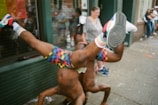 Two people are performing acrobatic dance moves on a sidewalk, with colorful outfits and socks. There is a small crowd gathering in the background, looking amused and engaged. A flag with bright colors is visible behind them.