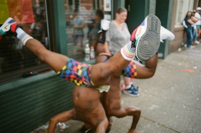 Two people are performing acrobatic dance moves on a sidewalk, with colorful outfits and socks. There is a small crowd gathering in the background, looking amused and engaged. A flag with bright colors is visible behind them.