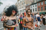 A lively scene on a city street, featuring a person in bold, theatrical attire with a large black headpiece and red sunglasses. Another person stands confidently beside them, wearing a harness and sunglasses, playfully sticking out their tongue. The street is bustling with people participating in a parade or festival, adorned with rainbow flags, adding to the festive atmosphere. Historic buildings line the background, adding an urban feel to the vibrant celebration.