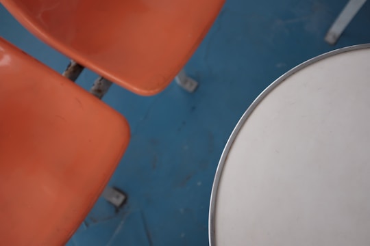Two orange plastic chairs are positioned next to a round, white tabletop with a metallic edge, set against a blue floor.