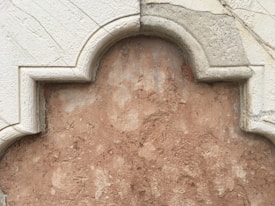 A detailed view of a textured wall, featuring a decorative architectural element with scalloped edges. The surface has a rough, earthy appearance with two contrasting sections; the border has a lighter, smoother finish compared to the central area which is more rugged and darker in tone.