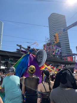 A colorful parade event with a large crowd in a city, featuring a person in a vibrant, multi-colored dress resembling a peacock's tail on a float. The environment is lively with a clear blue sky above and tall buildings surrounding the area.
