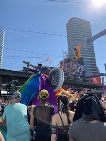 A colorful parade event with a large crowd in a city, featuring a person in a vibrant, multi-colored dress resembling a peacock's tail on a float. The environment is lively with a clear blue sky above and tall buildings surrounding the area.