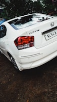 A white Honda City car is parked on a dirt surface. The back of the car is visible, showing the tail lights and license plate. There is greenery and trees in the background.
