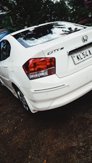 A white Honda City car is parked on a dirt surface. The back of the car is visible, showing the tail lights and license plate. There is greenery and trees in the background.