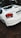 A white Honda City car is parked on a dirt surface. The back of the car is visible, showing the tail lights and license plate. There is greenery and trees in the background.