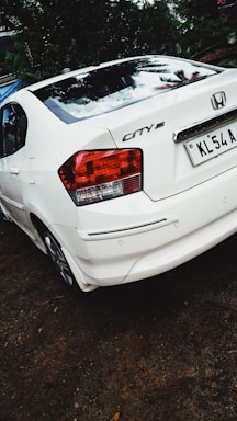 A white Honda City car is parked on a dirt surface. The back of the car is visible, showing the tail lights and license plate. There is greenery and trees in the background.