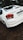 A white Honda City car is parked on a dirt surface. The back of the car is visible, showing the tail lights and license plate. There is greenery and trees in the background.