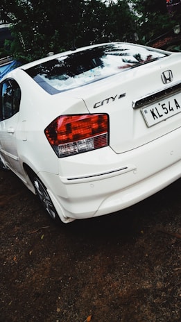 A white Honda City car is parked on a dirt surface. The back of the car is visible, showing the tail lights and license plate. There is greenery and trees in the background.