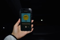 A hand is holding a smartphone displaying the song 'Be Nice' by The Black Eyed Peas featuring Snoop Dogg. The phone shows a yellow smiley face on the album cover against a blue background. The setting is dark, possibly inside a car, with some faint lights visible in the background.