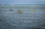 Several kayakers are paddling on a calm body of water. The scene includes multiple groups of people in brightly colored kayaks, wearing life jackets and enjoying the outdoor activity. In the background, there are other boats, including a sailboat and a passenger boat.