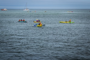 Several kayakers are paddling on a calm body of water. The scene includes multiple groups of people in brightly colored kayaks, wearing life jackets and enjoying the outdoor activity. In the background, there are other boats, including a sailboat and a passenger boat.