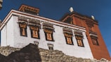A traditional Tibetan-style building with intricate window frames and a combination of white and red exterior walls. The structure is surrounded by rocky terrain and set against a clear blue sky.
