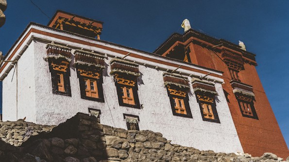 A traditional Tibetan-style building with intricate window frames and a combination of white and red exterior walls. The structure is surrounded by rocky terrain and set against a clear blue sky.