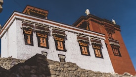 A traditional Tibetan-style building with intricate window frames and a combination of white and red exterior walls. The structure is surrounded by rocky terrain and set against a clear blue sky.